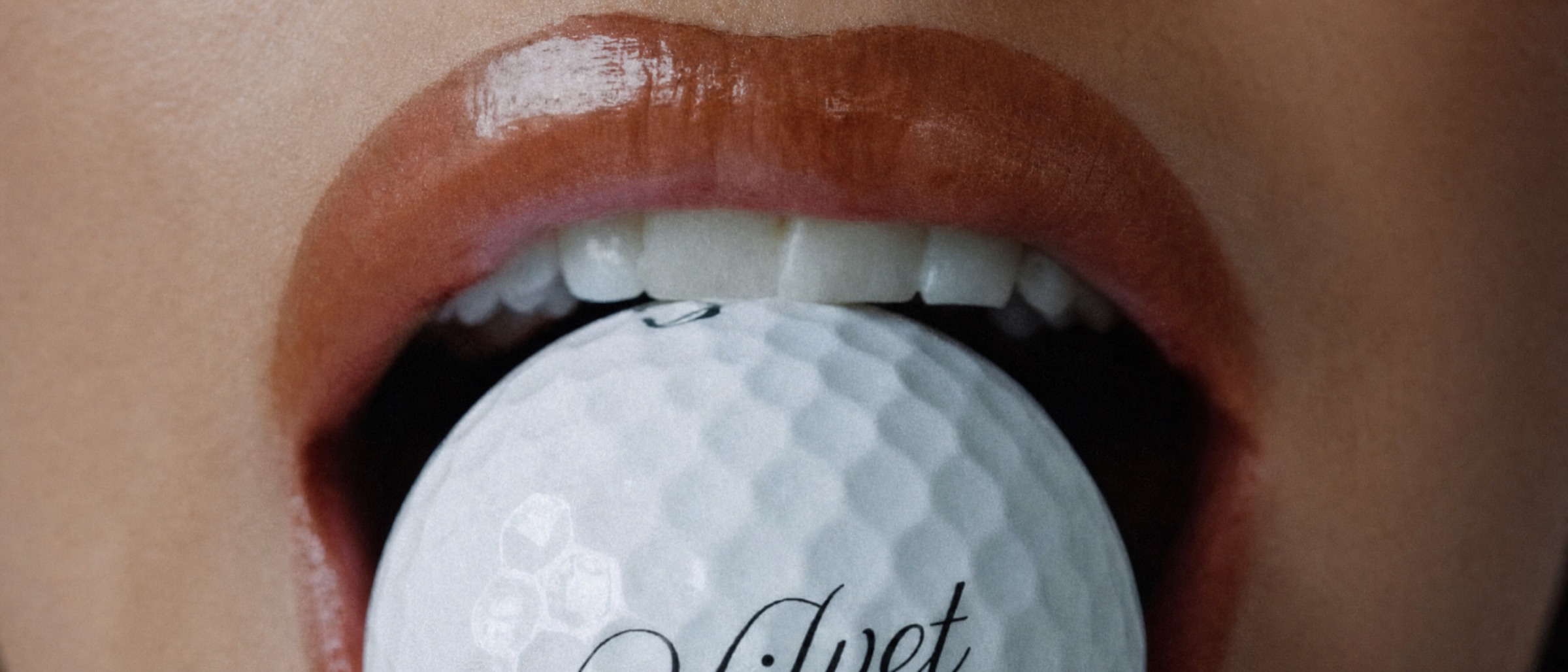 Close crop of a golf ball held between glossed lips.