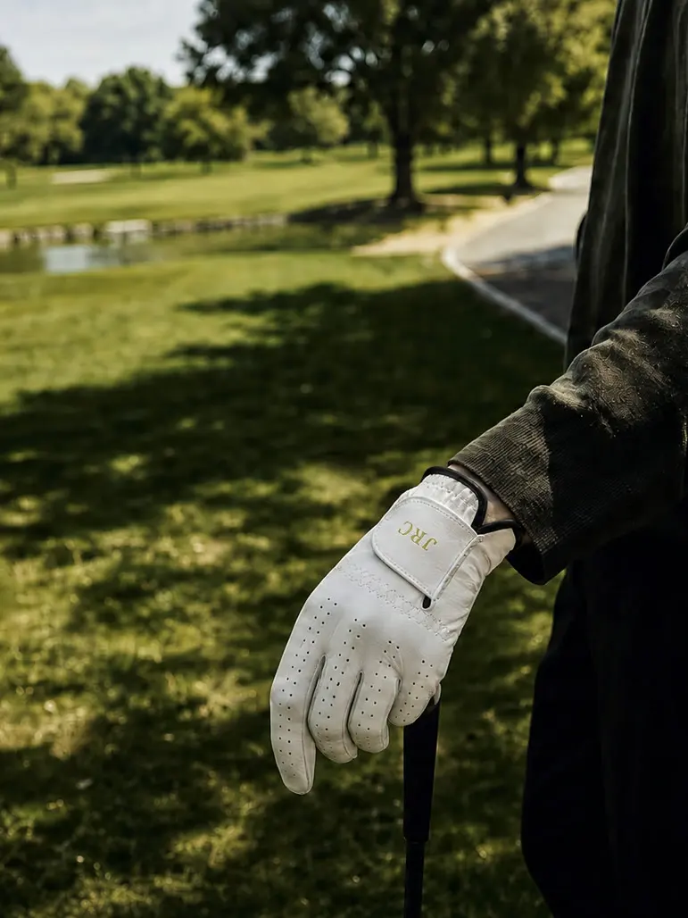 A golfer resting a gloved hand on his club by the fairway, initials JRC on the strap.